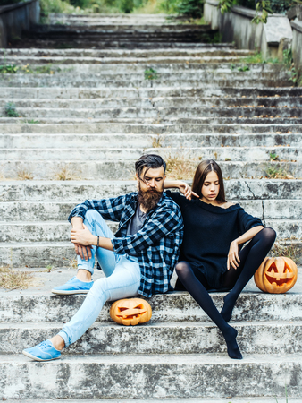 young halloween couple of bearded man with beard and mustache and girl in black tights sit on stony stairs with traditional autumn holiday symbol of orange spooky pumpkin outdoorの写真素材