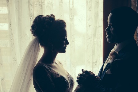 Silhouette of bride beautiful young woman and groom handsome african American man with wedding bouquet look at each other with love on background of windowの写真素材
