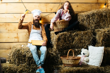 Angry man chef cook or baker with beard and moustache in hat toque and cute girl cookee teenager in apron sit on straw bales on rustic backgroundの写真素材