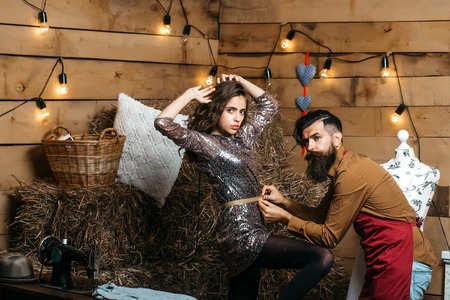 Bearded man tailor or dressmaker takes measurements from pretty girl customer or sexy model with curly brunette hair in rustic workshopの写真素材
