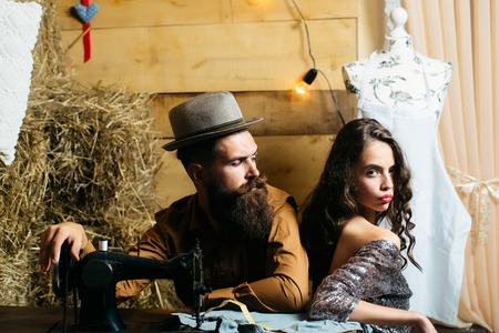 Bearded man tailor or dressmaker in hat and pretty girl customer or sexy model with curly brunette hair and undressed back sit near vintage sewing machine in rustic workshopの写真素材