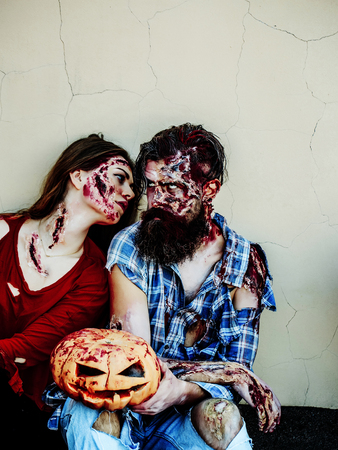 Zombie couple of bearded man hipster or war soldier and bloody young woman with wounds and red blood with Halloween pumpkin sit on beige wallの写真素材