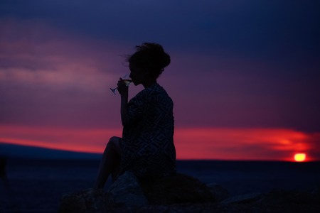 pretty sexy female silhouette of woman or girl with wine glass outdoor over dramatic dark sky with clouds and sea or ocean water horizon on evening or twilight natural sunset backgroundの写真素材