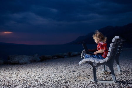 cute baby boy child with laptop or computer on bench outdoor over dark twilight sky with clouds on beach with sea or ocean water on evening natural background, copy spaceの写真素材