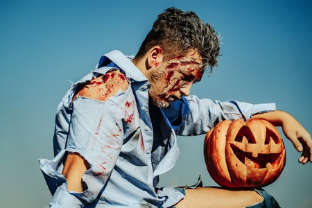 Handsome zombie man bloody young male or war soldier with wounds and red blood with Halloween pumpkin outdoors on blue skyの写真素材
