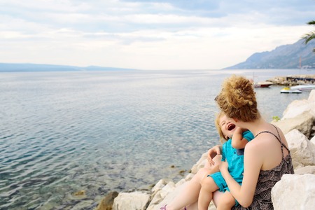 Happy mother and baby son sit on stones on blue sea beach and enjoy beautiful summer dayの写真素材