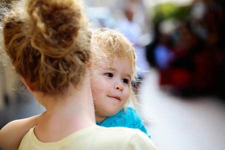 Mother young woman holds happy son baby boy with blonde hair in blue shirt sunny day outdoor in street on blurred backgroundの写真素材