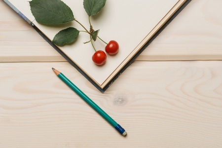 Two ripe red cherries with green leaves on open paper diary and graphite pencil on light wooden...