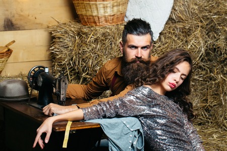 Bearded man tailor or dressmaker and pretty girl customer or sexy model with curly brunette hair in dress pose near vintage sewing machine in rustic workshopの写真素材