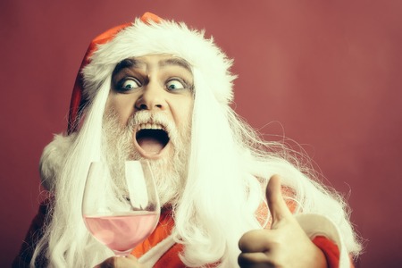 New year man with surprised face has long white beard and hair in red santa claus christmas coat and hat holding wine glass with liquid on studio background, copy spaceの写真素材