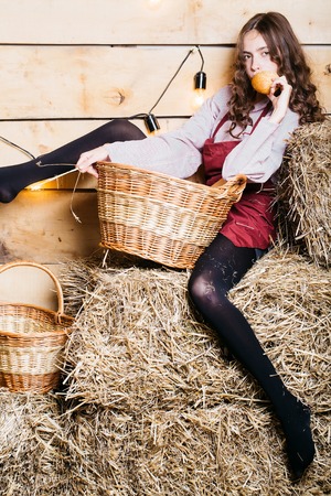 Pretty girl teenager young woman cookee helper in apron eats bun bread on straw bales with wicker basket on rustic backgroundの写真素材