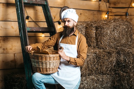 Handsome man chef cook or baker with beard and moustache in hat toque and apron stands on wooden ladder with basket on rustic backgroundの写真素材