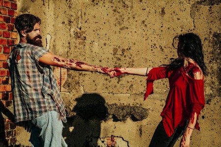 Halloween zombie couple of bearded man hipster or war soldier and bloody young woman with wounds and red blood hold hands outdoors on brick wallの写真素材