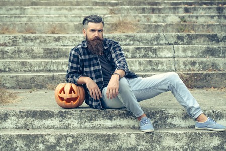 young halloween bearded man with beard and mustache in jeans and stylish checkered shirt sitting on stony stairs with traditional autumn holiday symbol of orange spooky pumpkin outdoorの写真素材