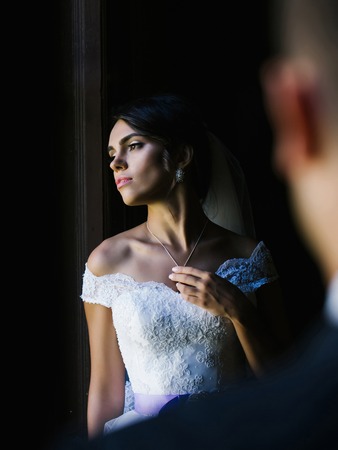 Sexy cute woman or girl bride with makeup on pretty face and veil in hair hold on necklace in white dress on dark backgroundの写真素材