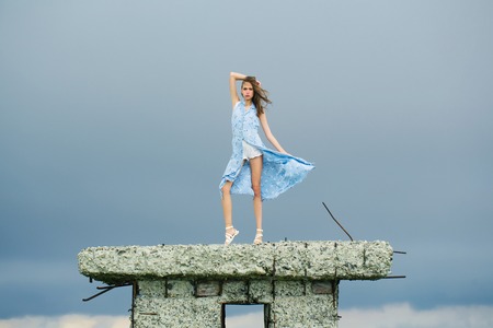 Pretty cute sensitive blonde woman or girl stands with raised hand in blue dress waving on wind outdoor on broken concrete bunkerの写真素材