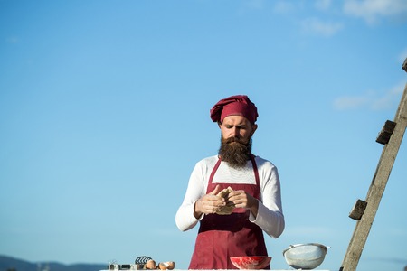 Man chef cook hipster with long beard on handsome face in red hat and apron cooking dough standing near ladder on blue sky background outdoorの写真素材