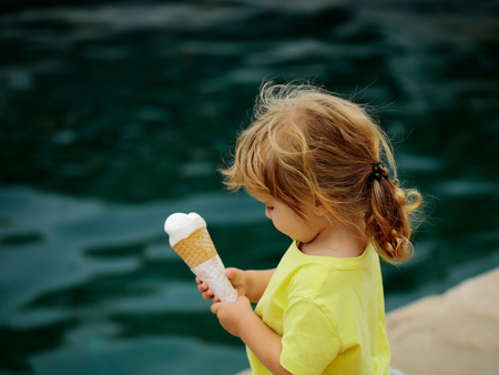 Cute baby boy child with curly blond hair ponytail in yellow shirt eats icecream outdoors on blue seaの写真素材