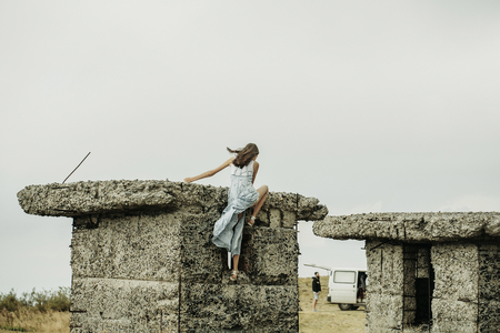 Pretty cute sensitive blonde woman or girl in blue dress waving on wind outdoor climbs on broken concrete bunkerの写真素材