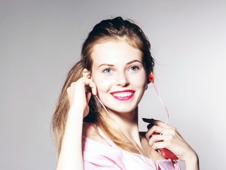 young sexy fashionable woman or girl with long brunette hair and red lips on pretty smiling happy face in stylish pink dress in studio on grey background with headset on musical playerの写真素材