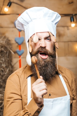 Brutal man chef cook or baker with beard and moustache in hat toque with wooden spoons and stirrers stands on straw bales on rustic backgroundの写真素材