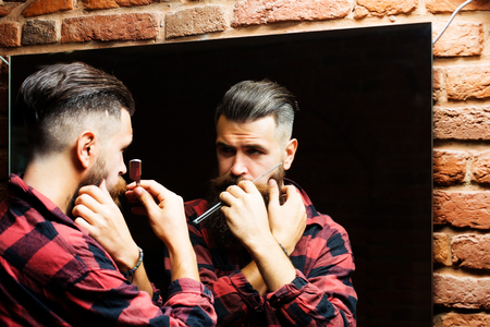 Handsome bearded man hipster with stylish haircut and beard holding old fashion razor in red checkered shirt looking at mirror with serious face in beauty salon or barbershop as barber near brick wallの写真素材