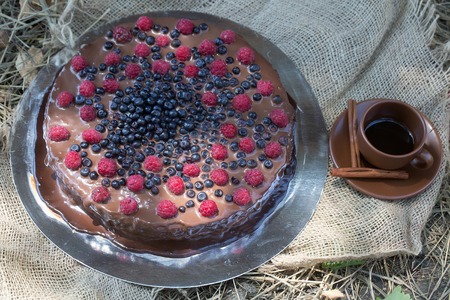 Chocolate cake dessert with fresh berries sweet blueberry and red raspberry on metallic plate and brown cup and saucer with coffee and aromatic pipes of cinnamon on sackcloth backgroundの写真素材