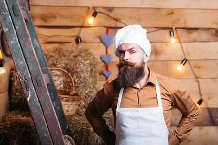 handsome bearded cook chef in white uniform and hat with long lush moustache on serious face standing near ladder with straw and lights in room on wooden backgroundの写真素材