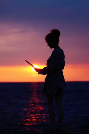 pretty sexy female silhouette of woman or girl with computer or laptop outdoor over dramatic dark sky with clouds and sea or ocean water horizon on evening or twilight on natural sunset backgroundの写真素材