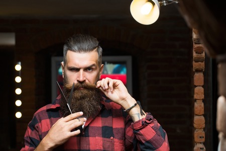 Handsome bearded man hipster with stylish haircut and beard holding scissors in red checkered shirt near mirror with serious face in beauty salon or barbershop as barber near brick wallの写真素材