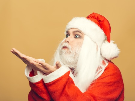 New year man with funny face with long white beard and hair in red santa claus christmas coat holding hands and blowing on yellow backgroundの写真素材