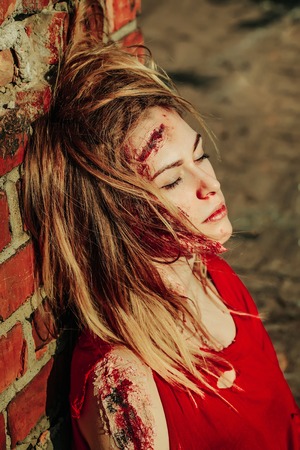 Halloween zombie girl pretty young woman with bloody blond hair with wounds and red blood sits outdoors at brick wallの写真素材