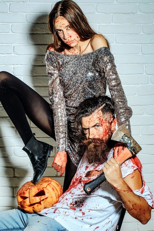 Zombie couple of bearded man hipster or war soldier with halloween pumpkin and axe and bloody young woman with wounds and red blood on brick wallの写真素材