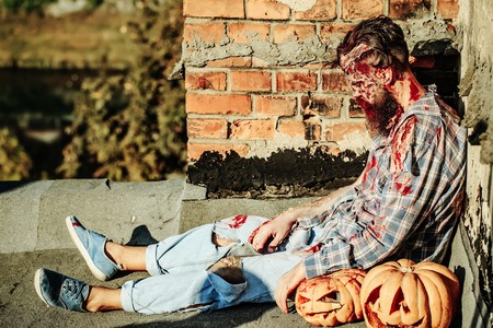 Bearded zombie man with beard creepy vampire or bloody war soldier with wounds and red blood sits with Halloween pumpkins outdoors on brick wallの写真素材