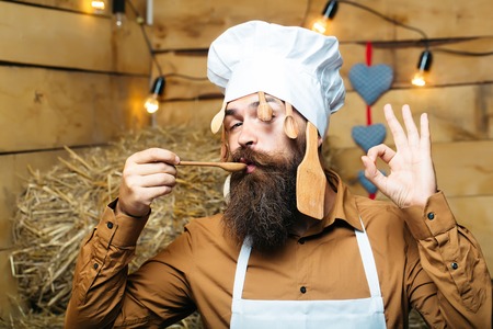 Funny man chef cook or baker with beard and moustache in hat toque with wooden spoons and ok hand gesture stands on straw bales on rustic backgroundの写真素材