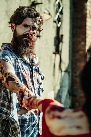 Halloween zombie couple of bearded man hipster or war soldier and bloody young woman with wounds and red blood hold hands outdoors on cement wallの写真素材