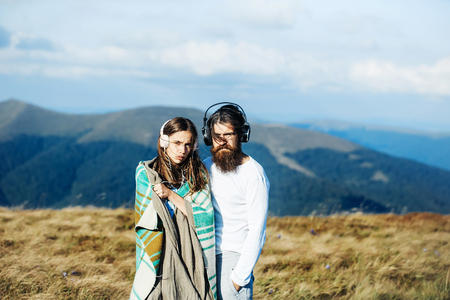 Young couple of pretty girl covered in blanket and bearded man hipster with beard listen to music with headphones on mountain sceneの写真素材