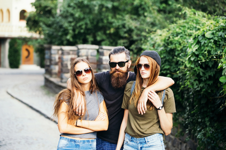 Tree friends handsome bearded hipster man hugging two pretty young girls in sunglasses casual clothes with serious faces standing on city street background outdoorの写真素材