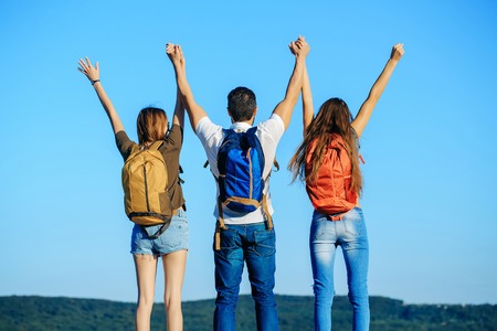 Bearded handsome man and two pretty sexy cute girls or women in jeans and shorts with backpacks outdoor on sunny day with raised hands on blue sky backgroundの写真素材