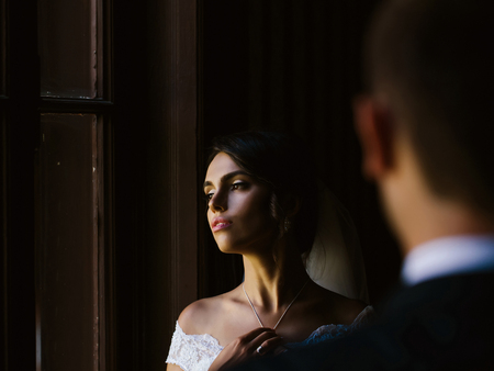 Sexy cute woman or girl bride with makeup on pretty face and veil in hair hold on necklace in white dress on dark backgroundの写真素材