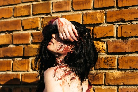 Halloween zombie girl pretty young woman with bloody brunette hair with wounds and red blood outdoors on brick wallの写真素材