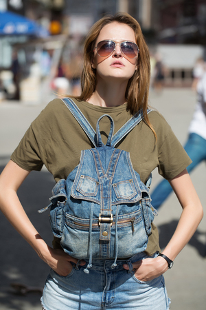 Young woman with pretty cute face in sunglasses in casual green shirt with denim backpack on breast holding her hands in jeans pockets on street background outdoorの写真素材