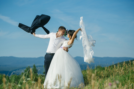 Handsome groom man shirt sleeve holds beautiful bride woman in long white wedding dress with flying veil in summer field on blue sky backgroundの写真素材