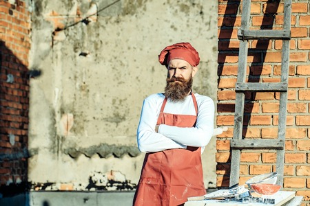 Man chef cook with bearded handsome serious face in red hat and apron with crossed hands on ladder...