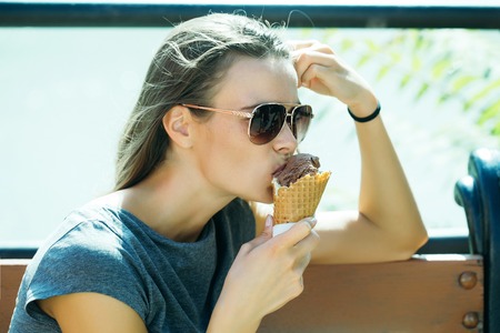 Young woman with pretty sexy cute face dark long hair in sunglasses dark casual shirt holding hand near head and eating tasty chocolate ice cream cornet outdoorの写真素材