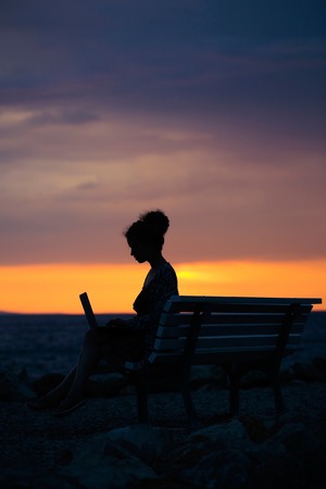 pretty sexy woman or girl with laptop or computer sits on bench outdoor over dark twilight sky with clouds on beach with sea or ocean water on evening natural backgroundの写真素材