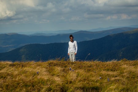 Handsome man bearded hipster with beard and moustache stands on mountain top on sunny summer day on cloudy skyの写真素材