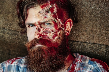 Bearded zombie man with beard halloween vampire or bloody war soldier with wounds and red blood outdoors on asphalt roadの写真素材