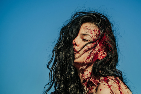 Halloween zombie girl pretty young woman with bloody brunette hair with wounds and red blood outdoors on blue skyの写真素材