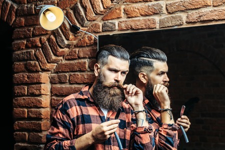 Handsome bearded man hipster with stylish haircut and beard holding old fashion razor in red checkered shirt near mirror with serious face in beauty salon or barbershop as barber near brick wallの写真素材
